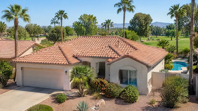 Early-1980s single-story stucco home in south Tempe, Arizona with a terracotta concrete tile roof, a backyard pool glimpsed beside the house, mature palms and a green park in the background