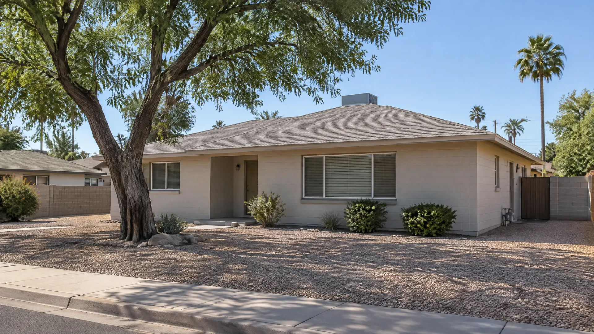 Small late-1940s early ranch-style home in an older Tempe, Arizona neighborhood with a low-pitched hip roof covered in new composition shingles, painted block walls, a mature shade tree and a gravel yard