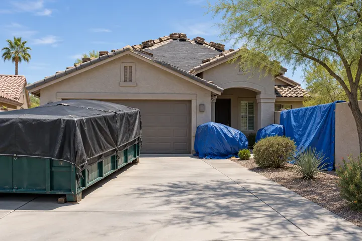 Tidy residential roofing job site in Arizona: a covered dumpster in the driveway, tarps protecting shrubs and a stucco wall, swept concrete, no people
